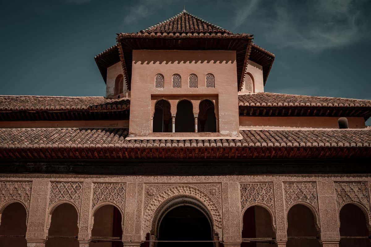 วิวภายนอกของส่วนหน้าอาคารที่ตกแต่งอย่างวิจิตรบรรจงของ Alhambra มีซุ้มโค้งแบบอิสลามที่ซับซ้อนและงานปูนปั้นที่ละเอียดประณีตใต้ท้องฟ้าสีฟ้า
