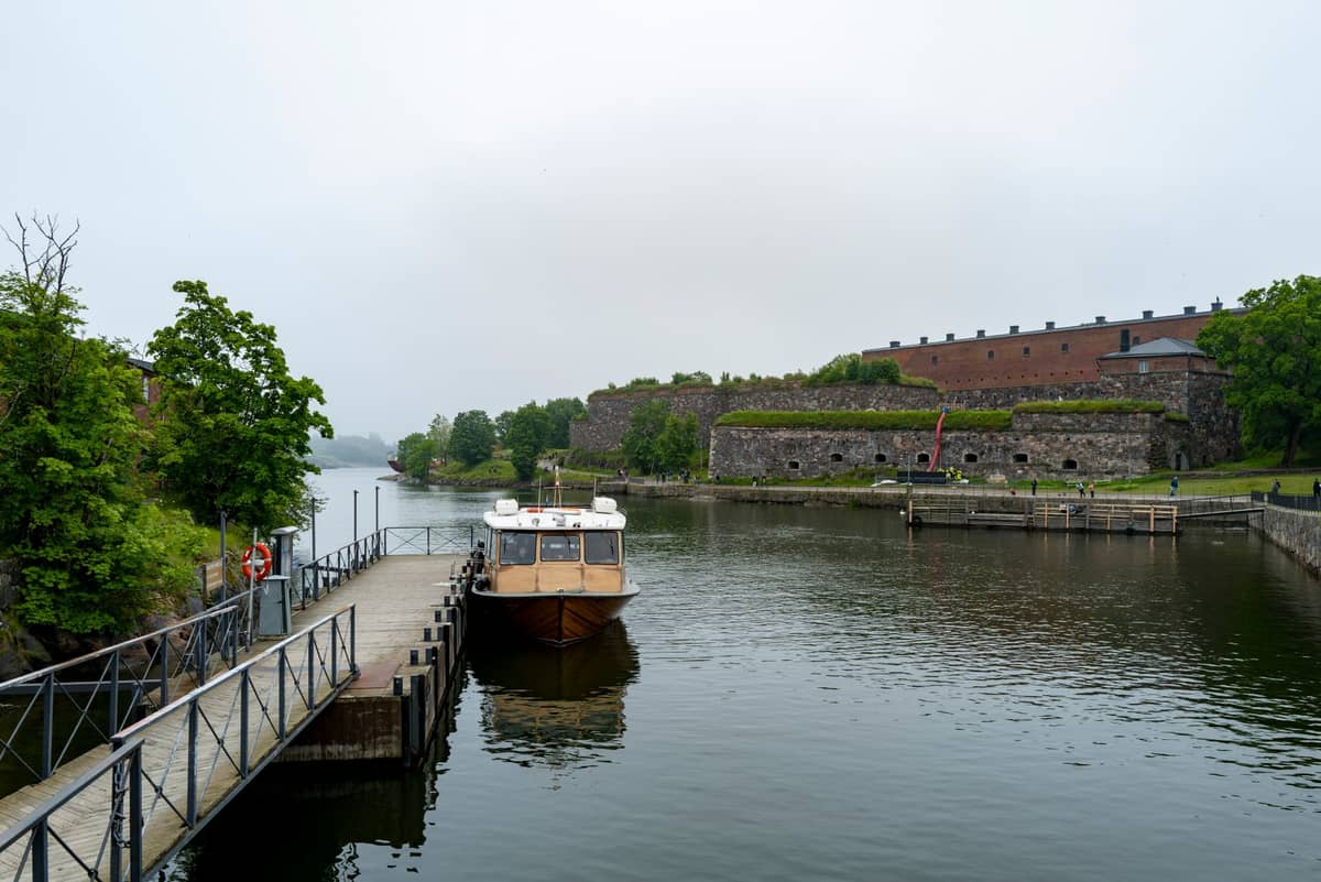 Pequena balsa atracada com as muralhas da fortaleza marítima de Suomenlinna e vegetação ao fundo num dia nublado em Helsínquia.