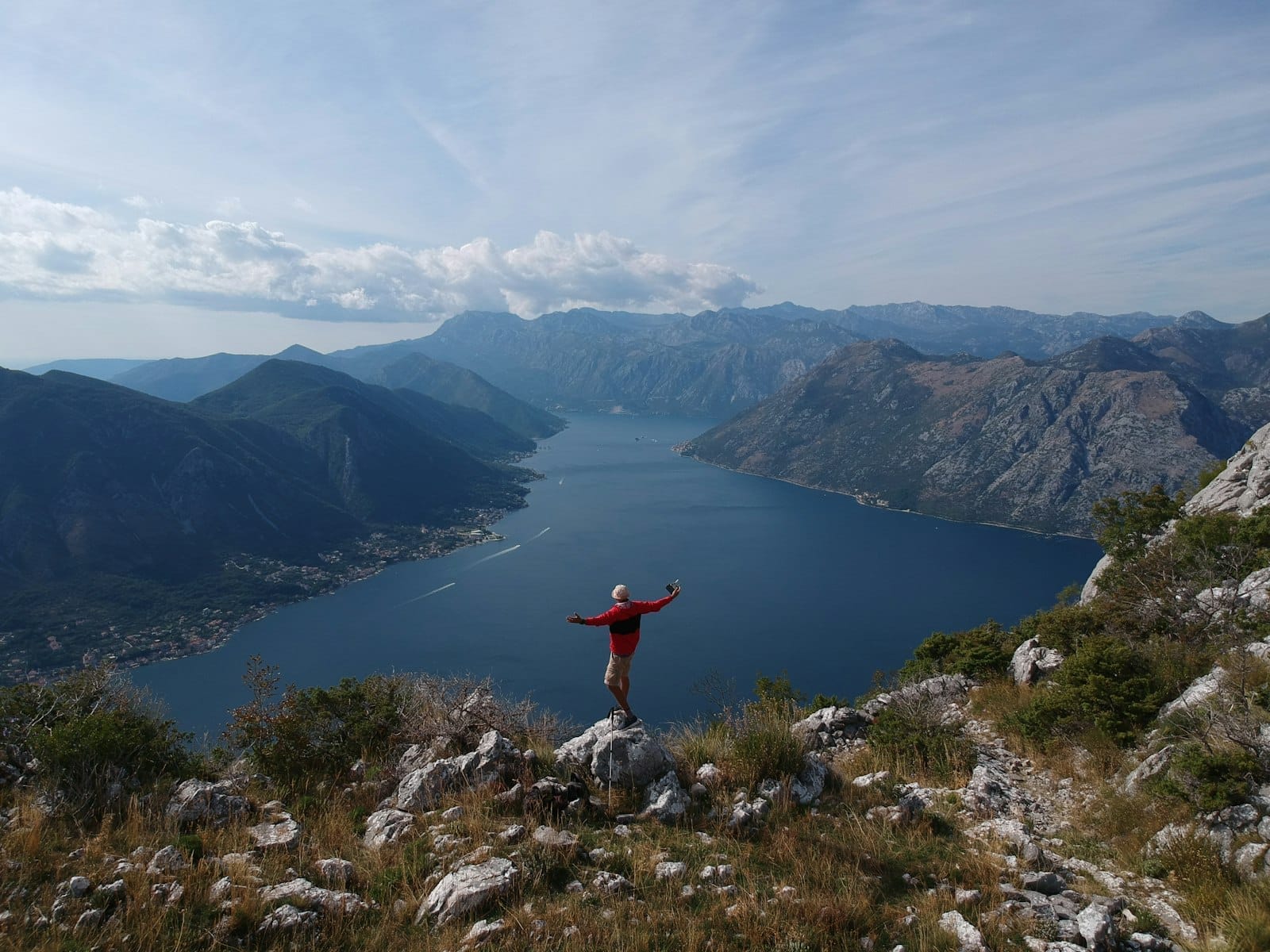 Vista da Baía de Kotor, Montenegro, com uma pessoa de jaqueta vermelha em pé sobre terreno rochoso contemplando a paisagem dramática.