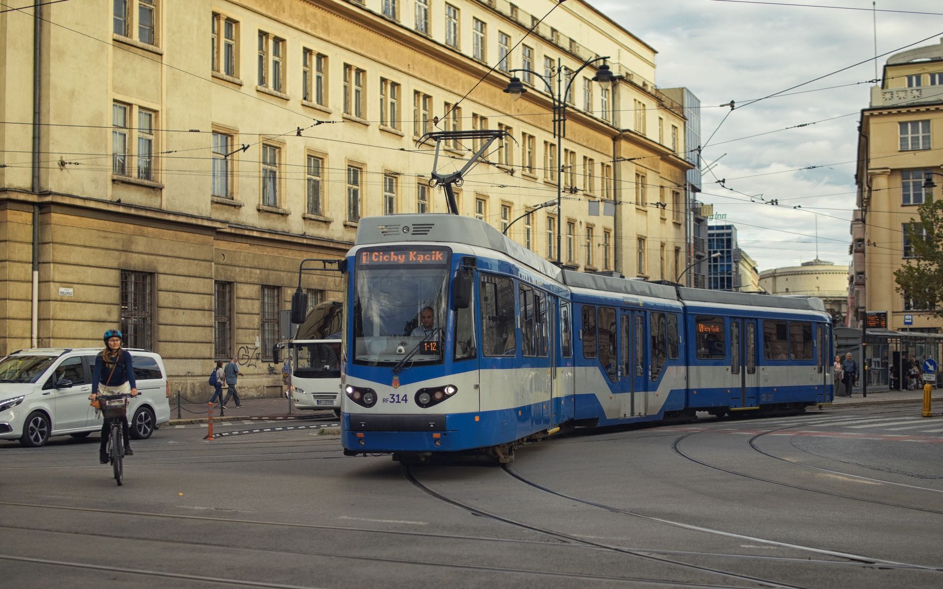 รถรางสมัยใหม่สีน้ำเงินและขาววิ่งไปตามถนนในเมือง Kraków มีผู้คนเดินและนักปั่นจักรยานอยู่ใกล้เคียงในเวลากลางวัน