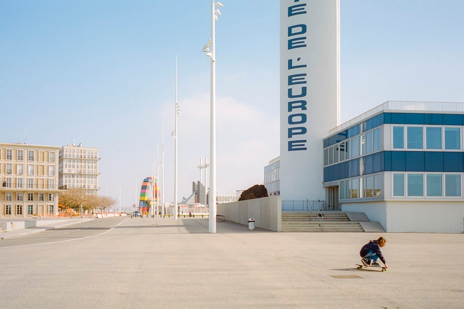 Vista ampia della Place de l'Europe a Le Havre con edifici modernisti, una colorata scultura pubblica e uno skateboarder sotto un cielo azzurro limpido.