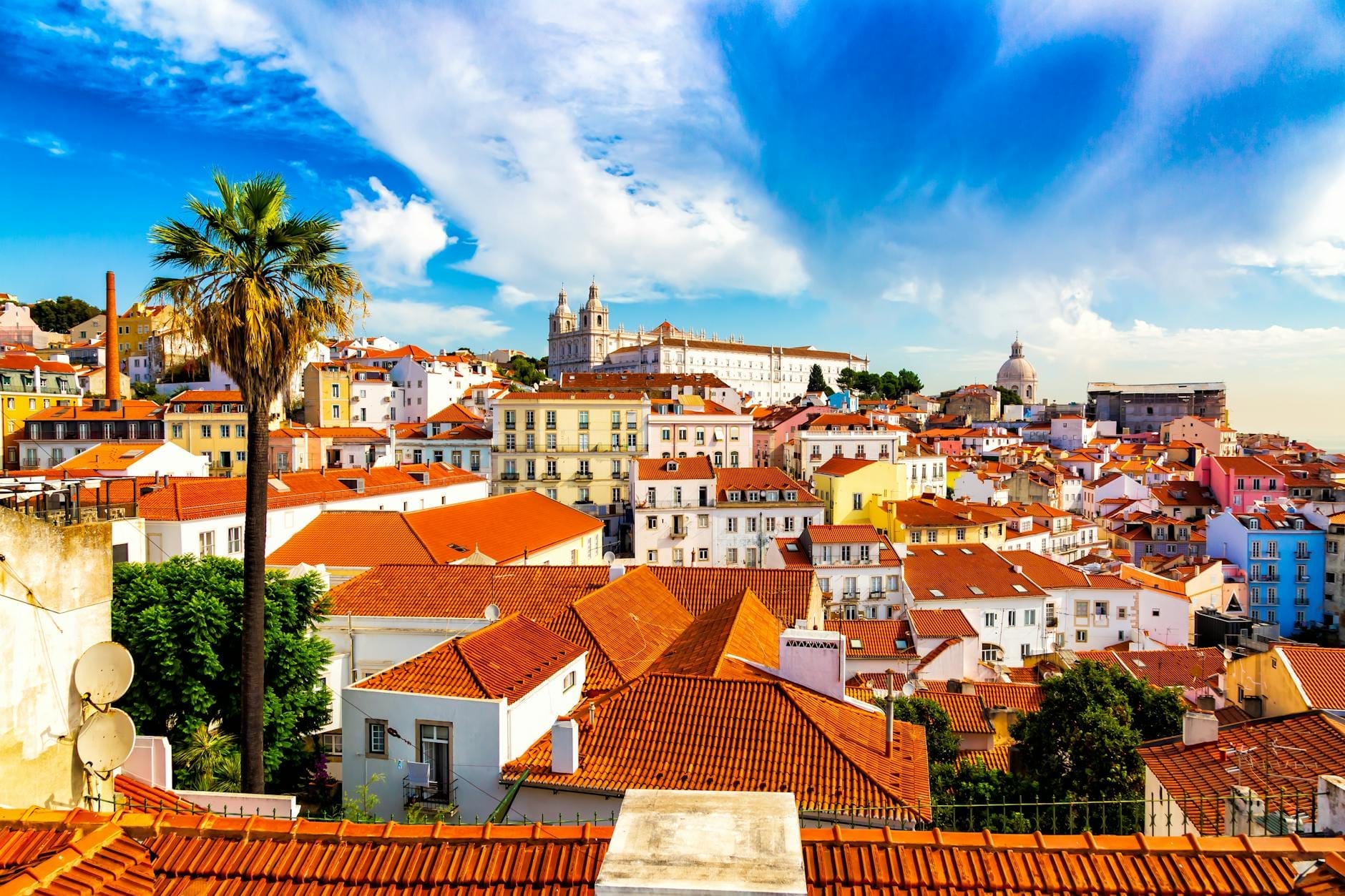 Vue panoramique lumineuse sur le quartier historique d'Alfama à Lisbonne avec des toits rouges, des bâtiments colorés, des palmiers et un ciel bleu spectaculaire par une journée ensoleillée.