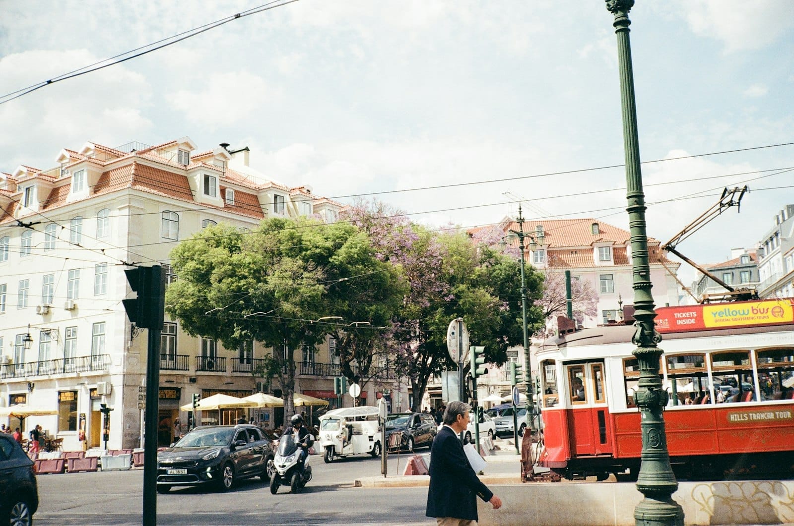 Tramway rouge classique de Lisbonne, voitures, motos et piétons dans une rue centrale animée avec des bâtiments aux couleurs pastel et des arbres en arrière-plan.