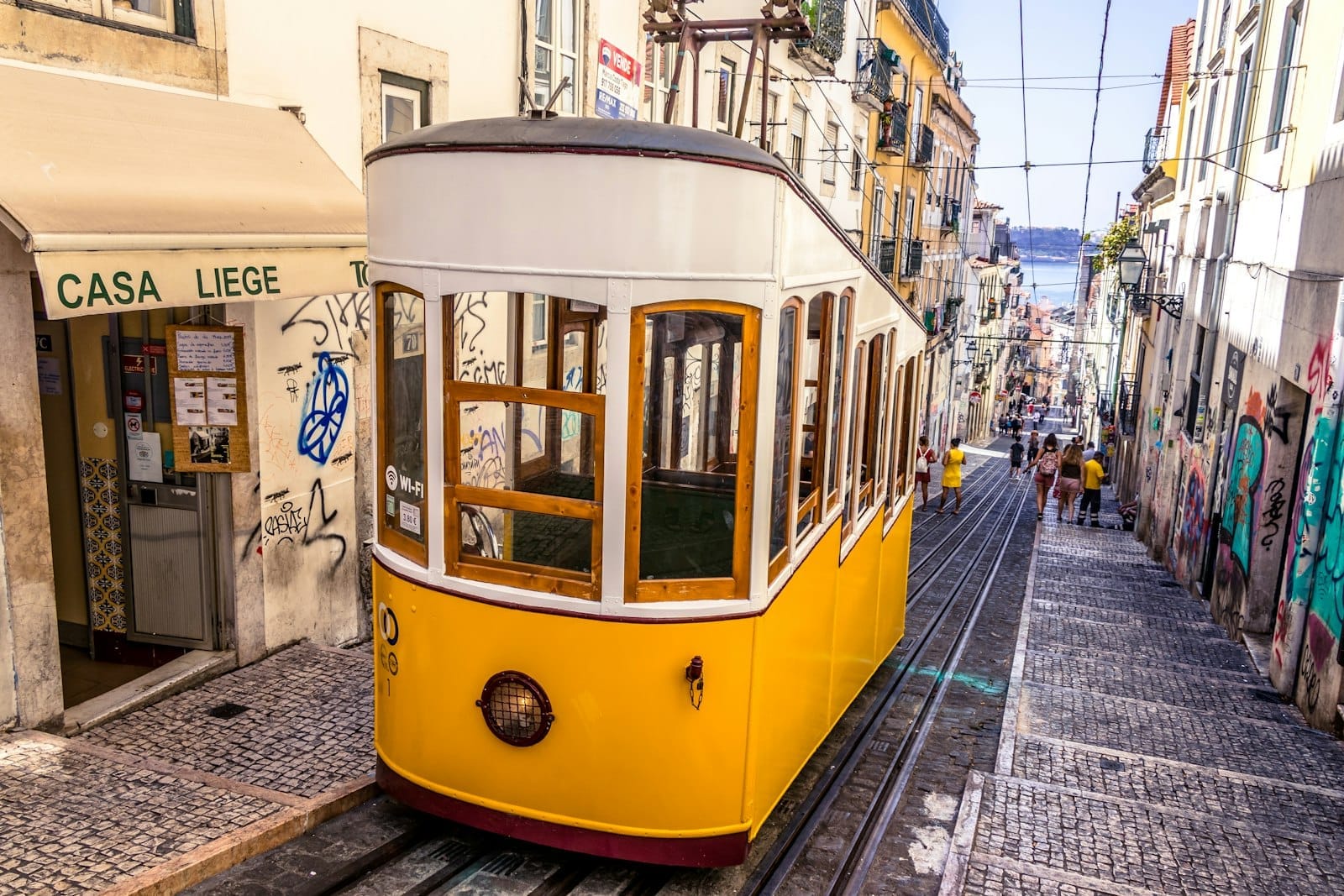 Tramway jaune classique de Lisbonne dans une rue pavée escarpée bordée de bâtiments colorés avec un aperçu du fleuve en arrière-plan.