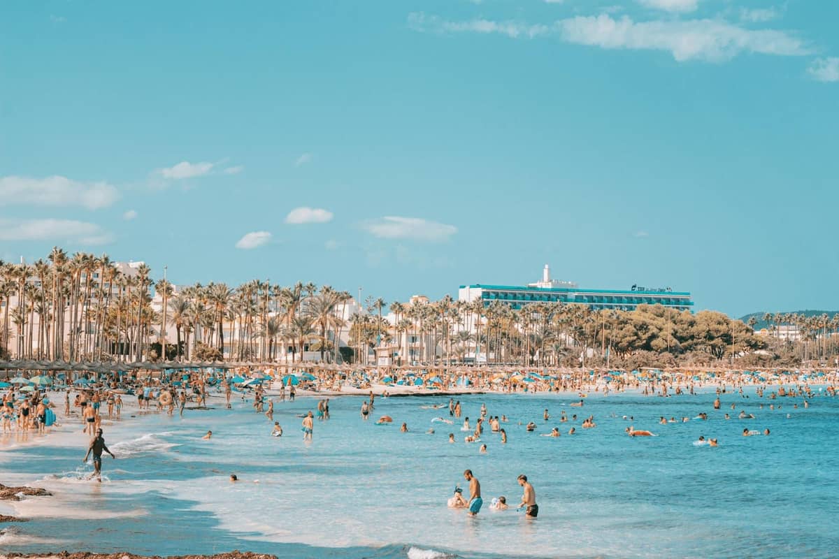 Movimentada praia de Mallorca com orla lotada, muitas pessoas nadando e tomando banho de sol, palmeiras e um grande hotel resort ao fundo.