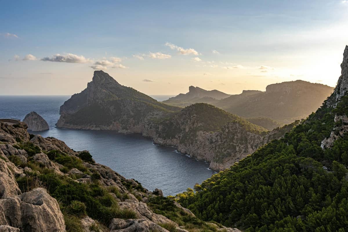 Vista costeira clara de Mallorca com falésias escarpadas, colinas verdejantes e mar calmo sob um céu luminoso, mostrando a variedade paisagística conforme a estação do ano.