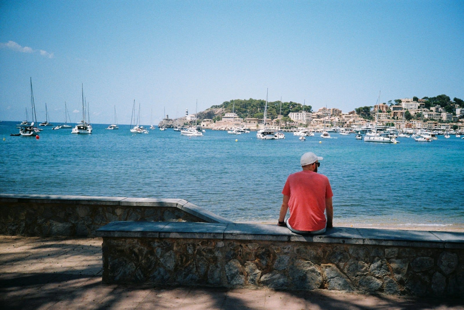 Vista da baía turquesa de Port de Sóller pontilhada de veleiros, edifícios mediterrâneos na encosta e um farol sob céus azuis limpos.