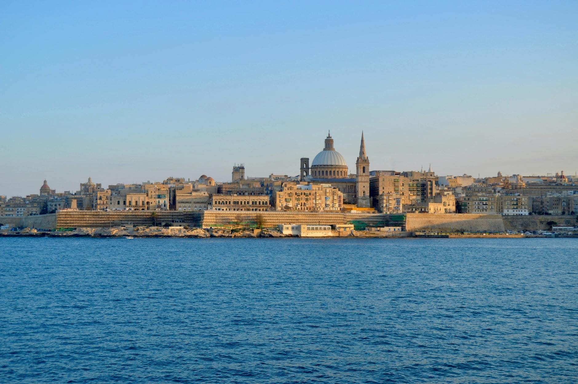 Vista do horizonte de Valletta, em Malta, com cúpulas históricas e torres de igrejas se erguendo sobre construções de pedra cor de mel, vistas de além das águas azuis do Mediterrâneo ao pôr do sol.
