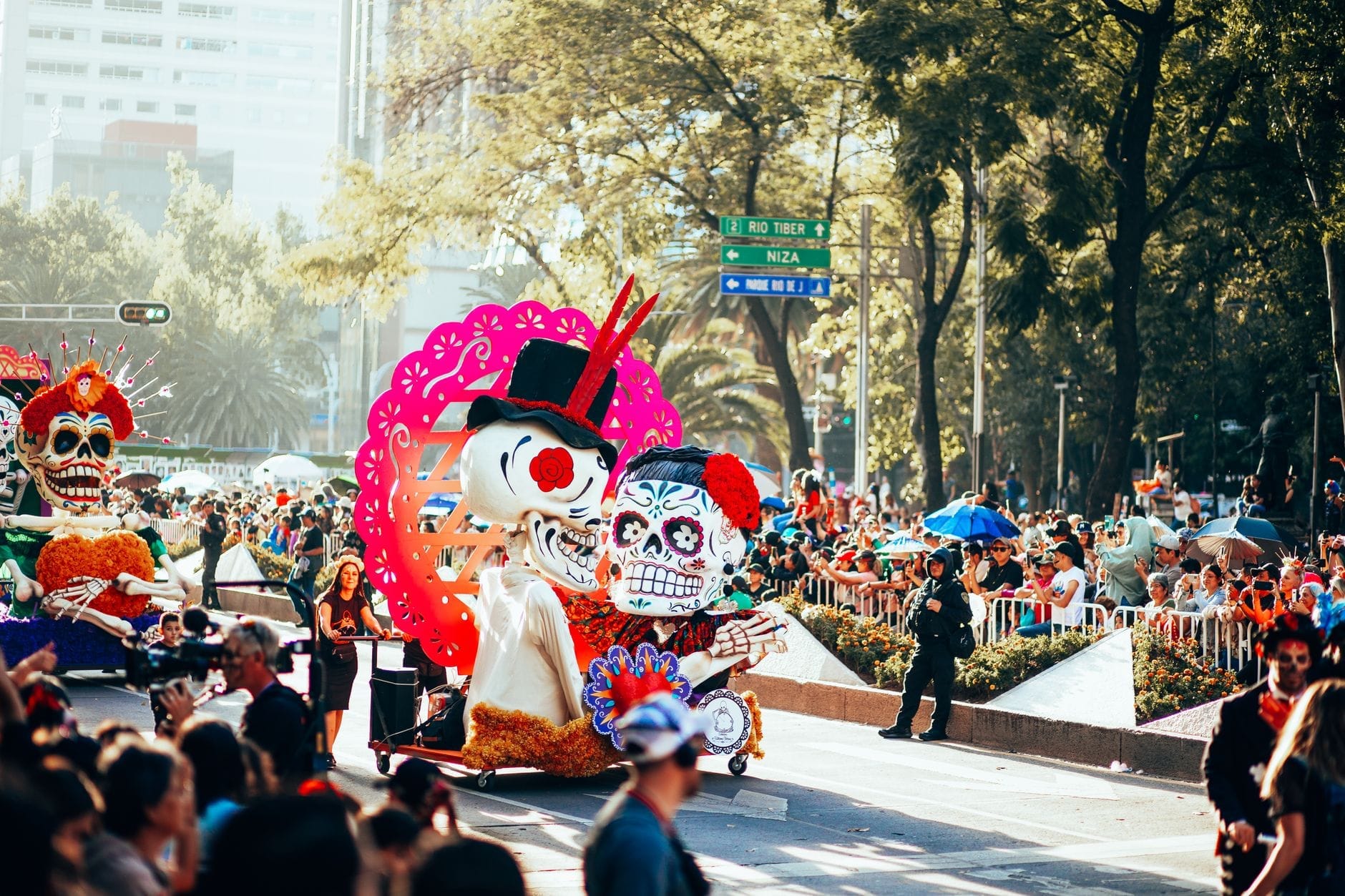 Desfile diurno durante o Día de Muertos na Cidade do México, com grandes carros alegóricos de calaveras e multidões densas ao longo das ruas.