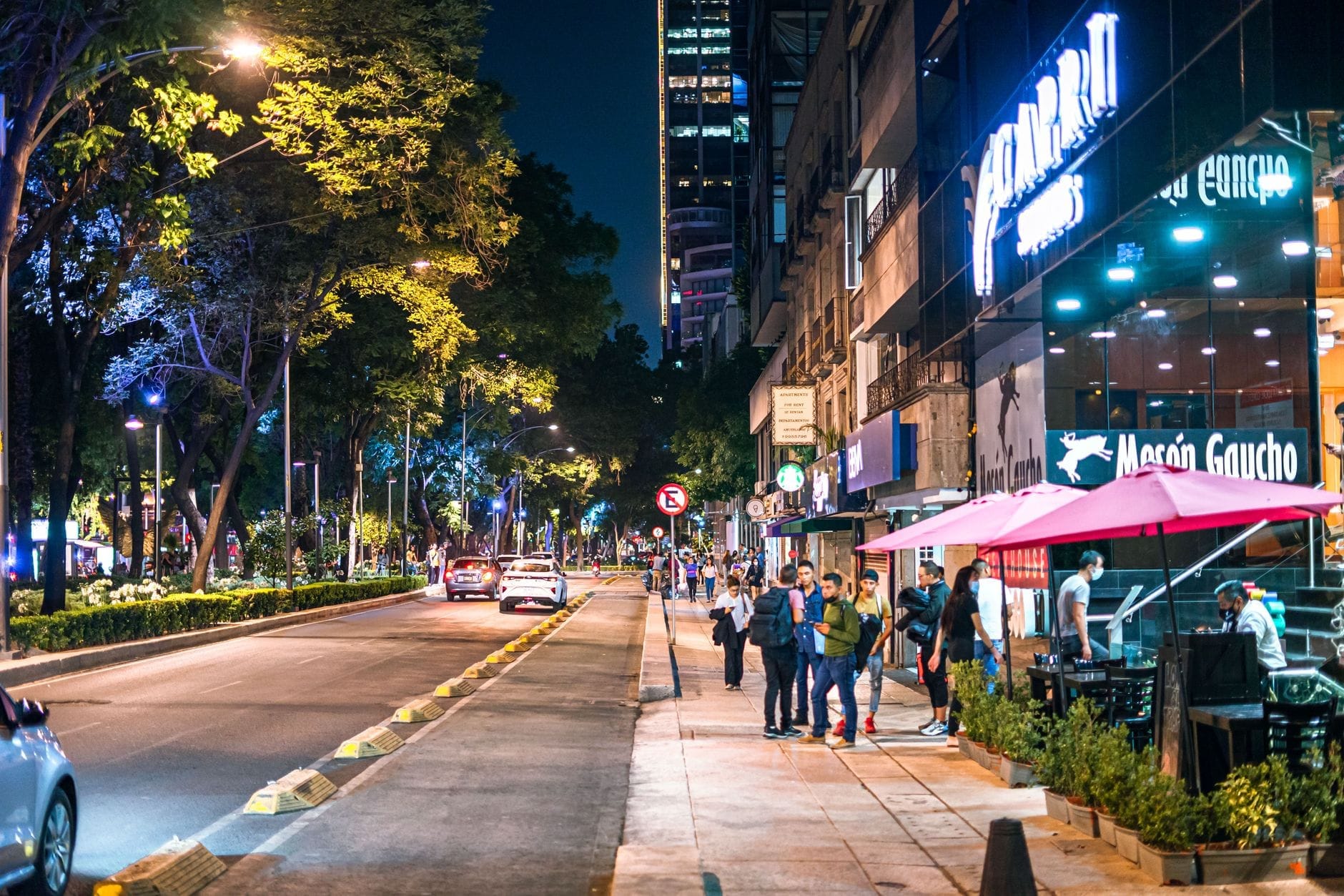 Rua bem iluminada da Cidade do México à noite com calçadas movimentadas, restaurantes, pessoas caminhando e carros passando, mostrando um bairro urbano agitado.