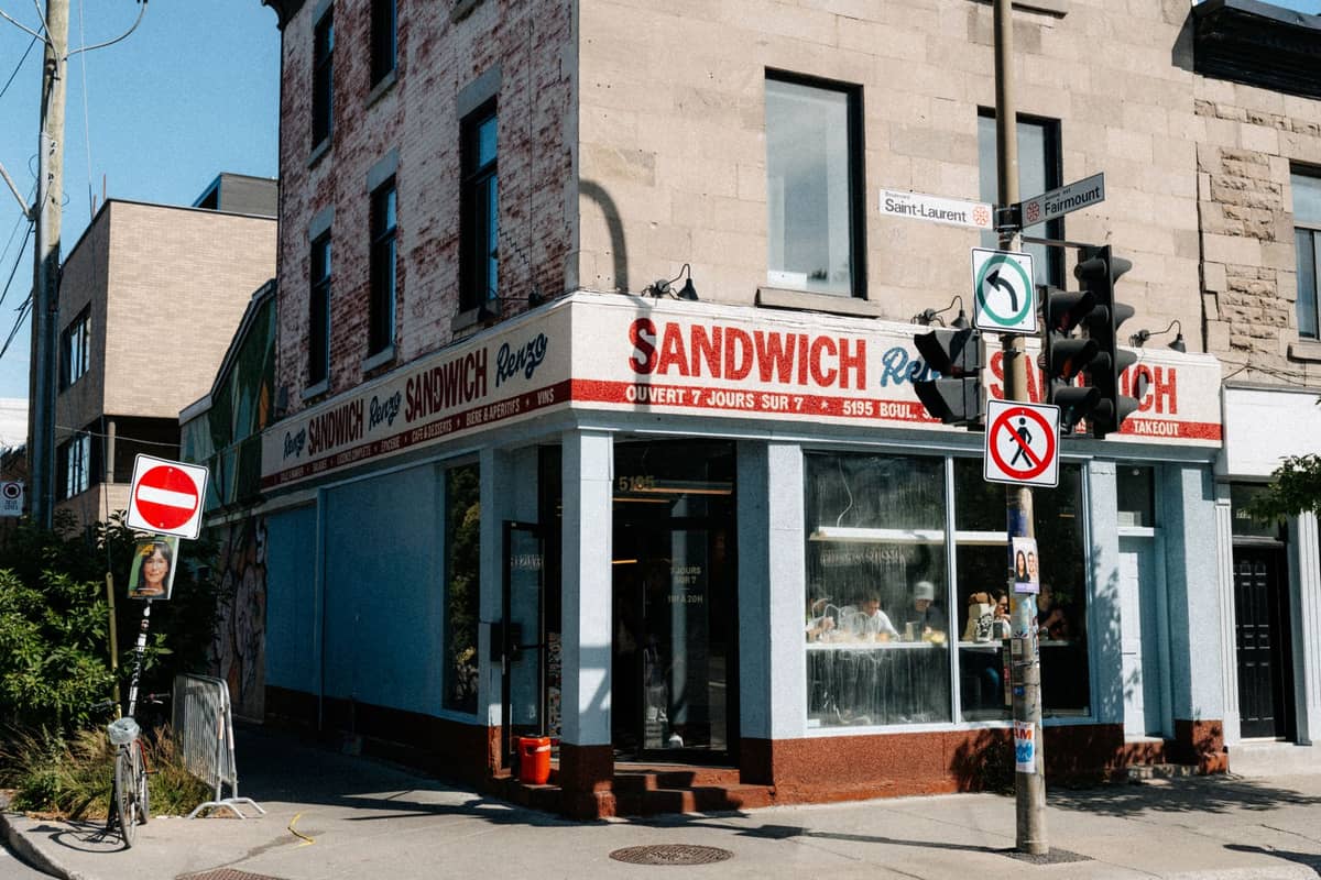 Zewnętrzna część klasycznego delikatesów w Montrealu na słonecznym rogu ulicy, z szyldami Sandwich i ludźmi w środku przy oknach.