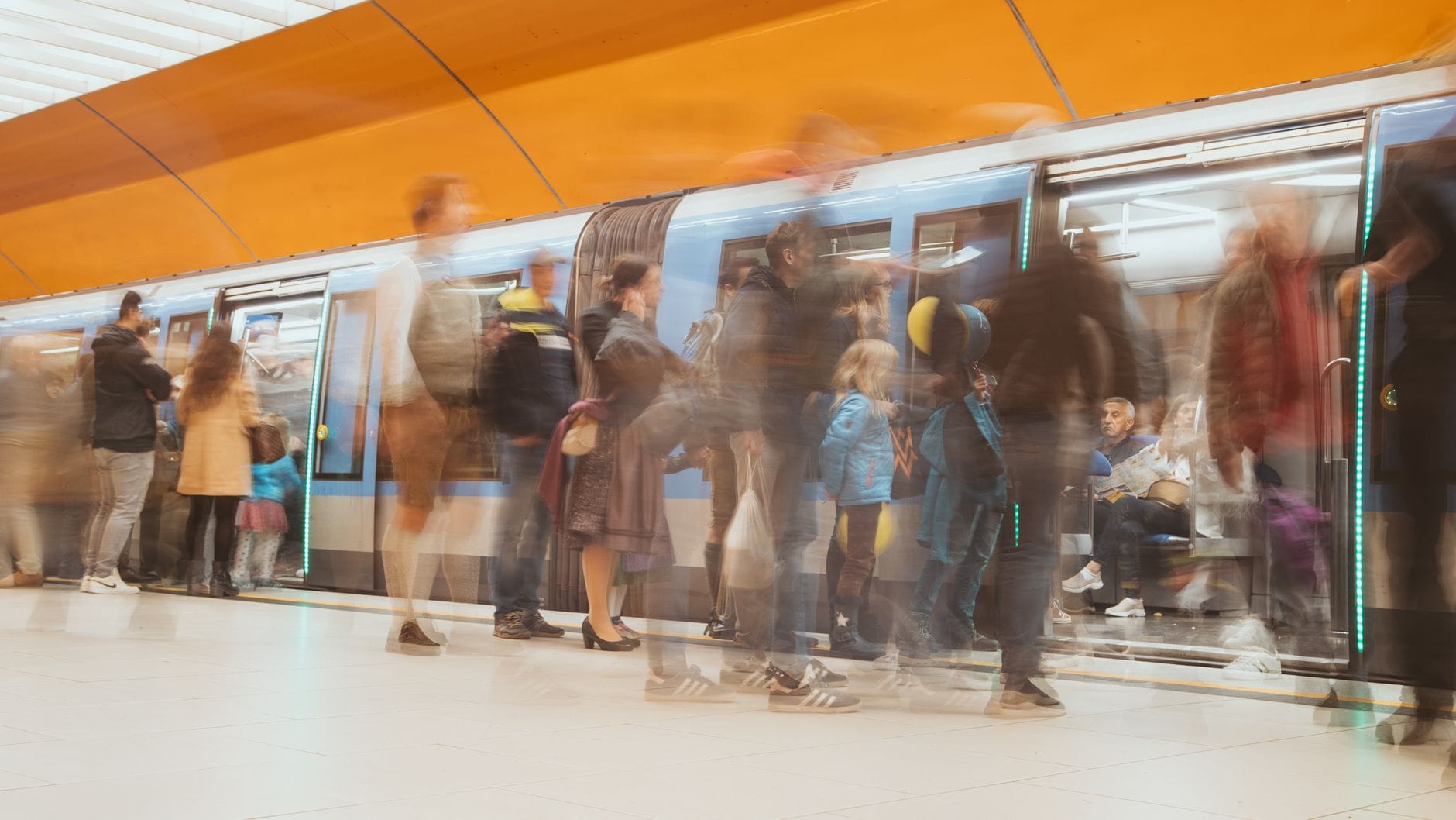 ฝูงชนผู้โดยสารกำลังขึ้นและลงจากรถไฟใต้ดิน U-Bahn สีน้ำเงินของ Munich ในสถานีใต้ดินสมัยใหม่ที่สว่างไสวพร้อมเพดานสีส้ม