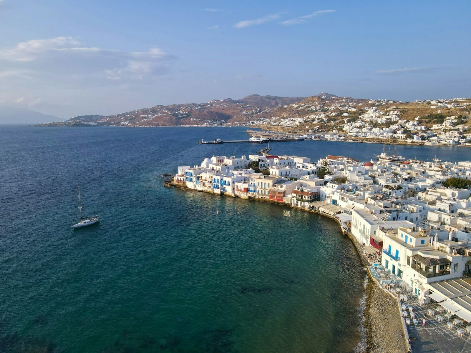 Vista aérea de la costa de Mykonos con edificios blancos, mar azul cristalino y un velero, mostrando la icónica arquitectura cicládica y el paisaje costero de la isla.