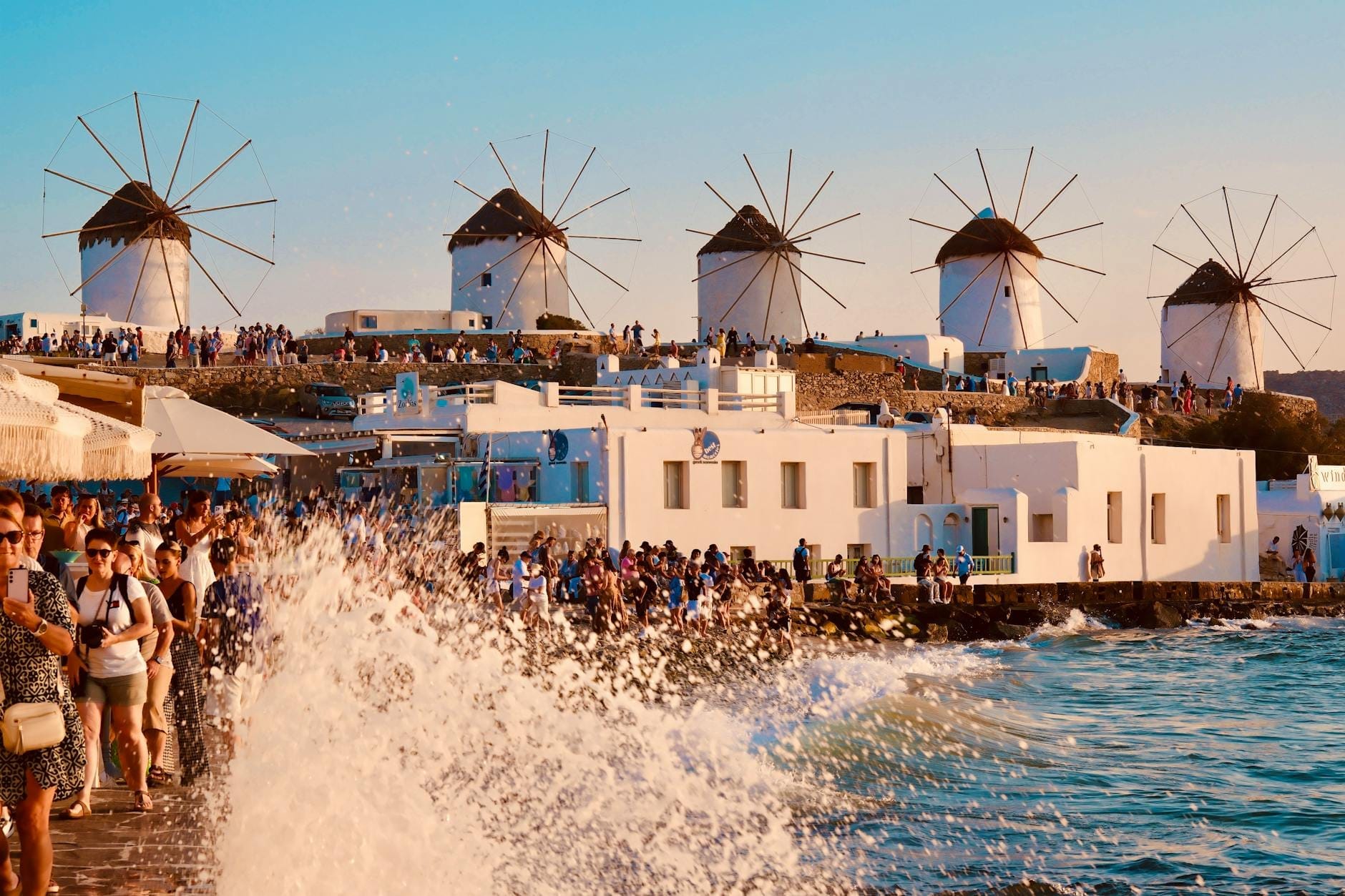 Multitudes de turistas pasean por el paseo marítimo de Mykonos con los icónicos molinos de viento al fondo, y las olas rompen contra la orilla en un día soleado.
