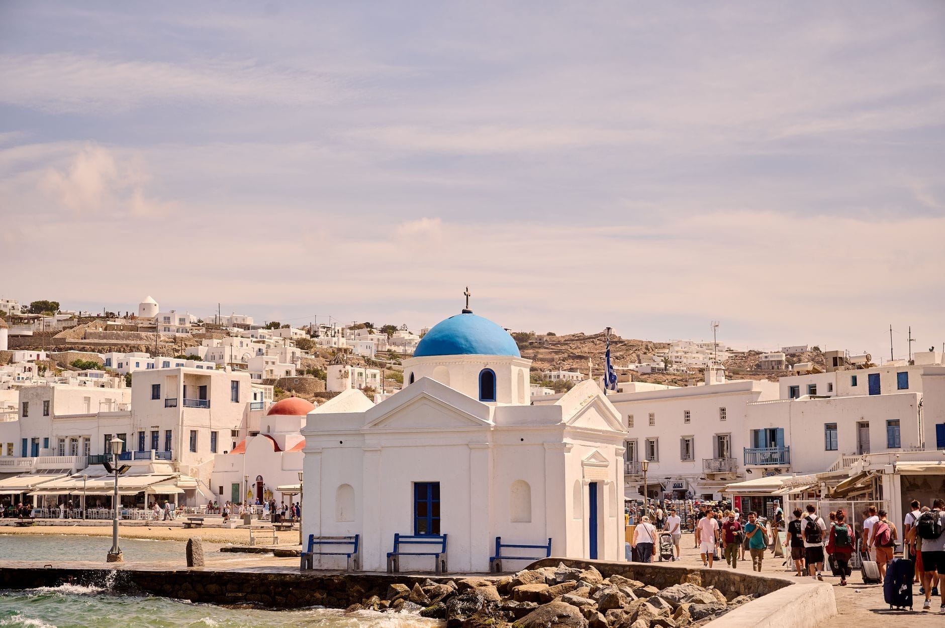 Multitudes de turistas y restaurantes frente al mar cerca de una iglesia blanca con cúpula azul en la ciudad de Mykonos, mostrando una animada y lujosa escena veraniega.
