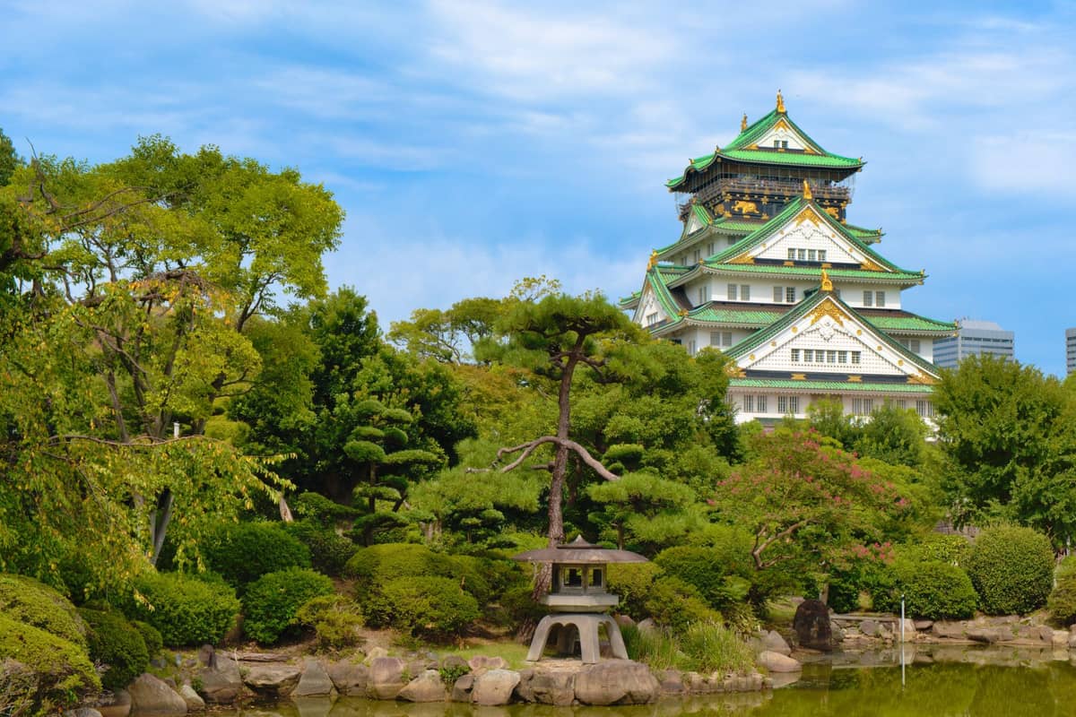 Osaka Castle framed by a lush Japanese garden, with the castle’s green roof and gold accents visible above trees and a pond in the foreground.