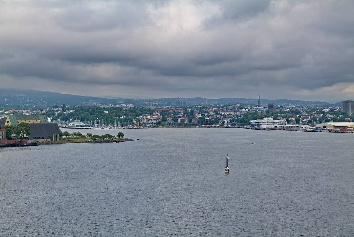 วิวกว้างของชายฝั่งออสโลพร้อมพิพิธภัณฑ์เรือไวกิ้ง Viking Ship Museum หลังคารูปสามเหลี่ยมอันโดดเด่นบนคาบสมุทร Bygdøy ท่าจอดเรือ และทิวทัศน์เมืองโดยรอบใต้ท้องฟ้ามีเมฆ