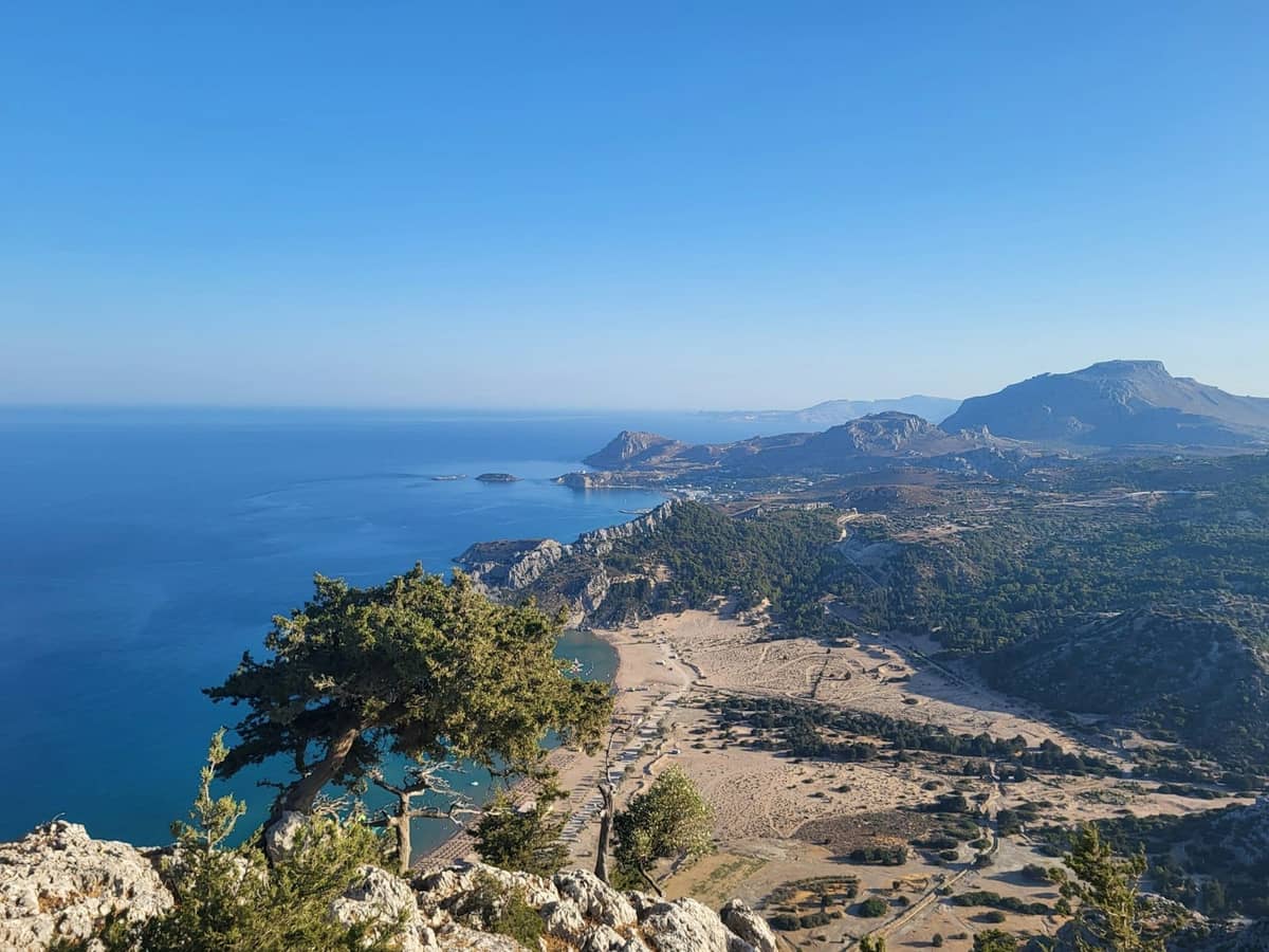 Elevated view of a wide sandy beach and turquoise waters on Rhodes, with hills and rugged coastline in the background under clear blue sky.