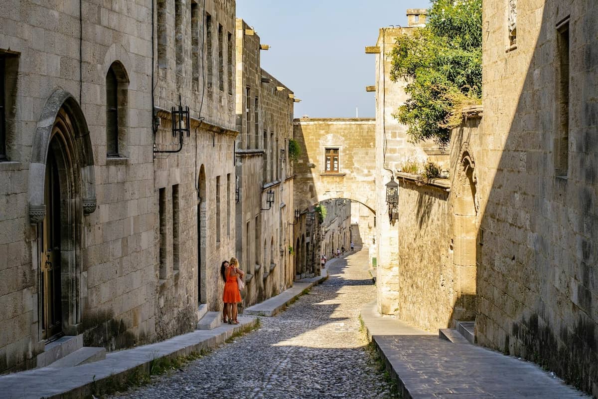 A medieval stone street in Rhodes Old Town with arched doorways, historic buildings, and two people walking in the distance.