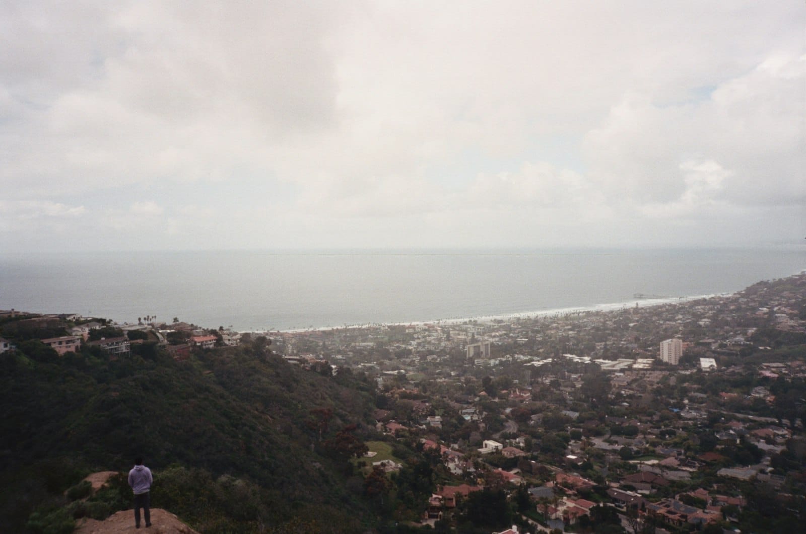 Vista panorâmica de uma cidade costeira com colinas, casas espalhadas e o mar se encontrando com o horizonte sob um céu nublado.