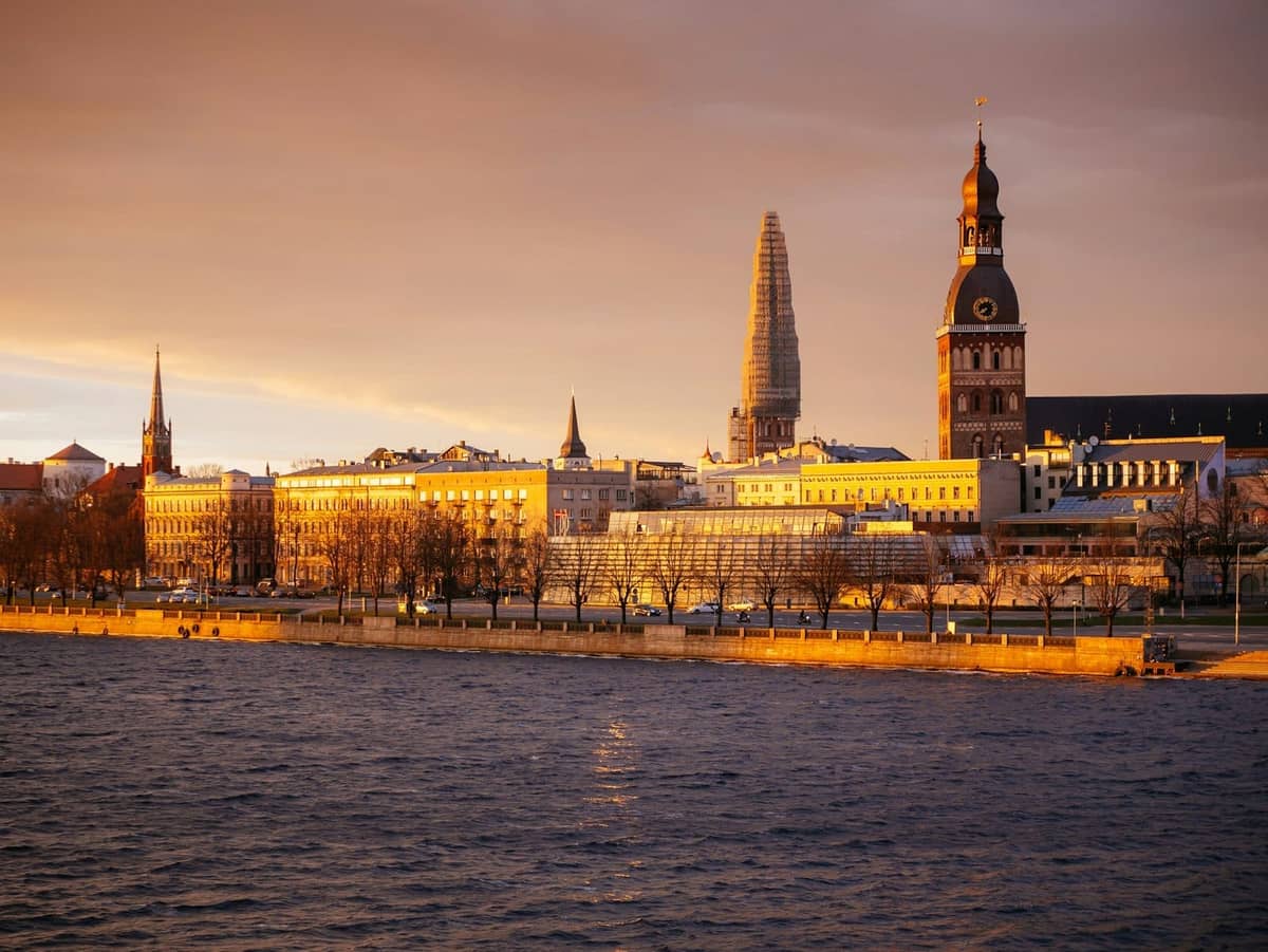Skyline der Rigaer Altstadt vom anderen Ufer der Daugava bei Sonnenuntergang, mit historischen Kirchturmspitzen und golden beleuchteten Gebäuden.