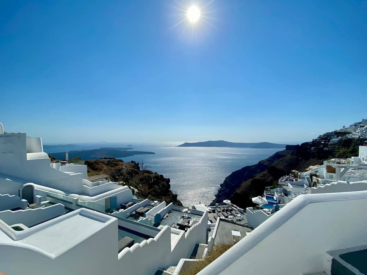 Sole brillante di mezzogiorno sulla caldera di Santorini con case imbiancate sul bordo della scogliera, mare azzurro e isole vulcaniche in lontananza.