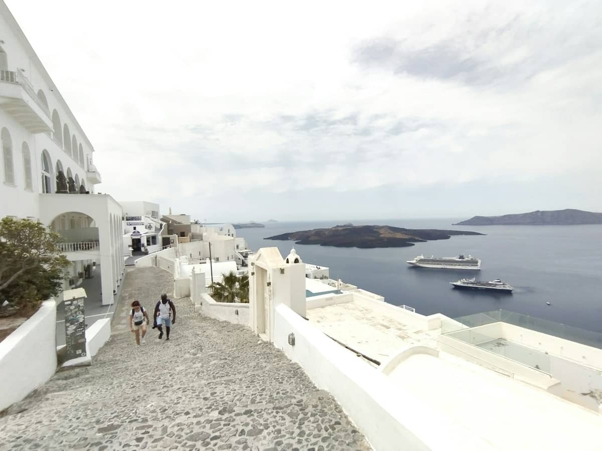 Due persone camminano su una tranquilla strada acciottolata e soleggiata a Santorini, con edifici bianchi, vista sul mare e navi da crociera visibili in basso.