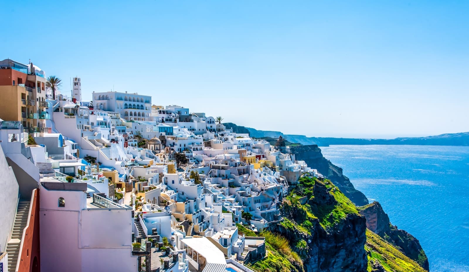 Casas e hotéis caiados de branco nos penhascos com vista para o azul profundo do Mar Egeu e da caldeira, sob um sol intenso e céu limpo, capturando a beleza clássica e a paisagem dramática de Santorini.