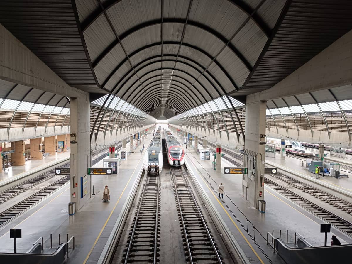 วิวกว้างของชานชาลาและรางรถไฟสมัยใหม่ภายในสถานีรถไฟ Santa Justa ในเมืองเซบิยา โดยมีรถไฟสองขบวนจอดรออยู่ใต้หลังคาโค้งที่โดดเด่น