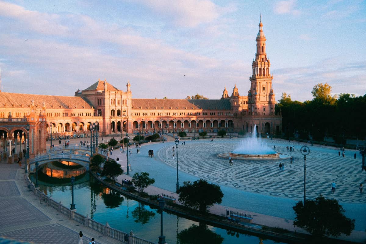 วิวกว้างของ Plaza de España ในเมือง Seville ช่วง Golden Hour พร้อมน้ำพุ คลอง สะพาน และผู้คนที่เดินอยู่รอบอาคารรูปครึ่งวงกลมอันยิ่งใหญ่