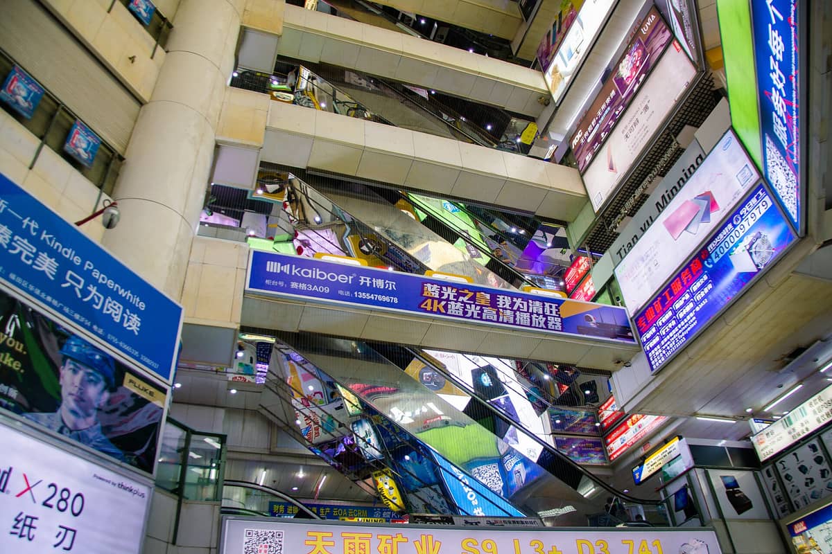 Vista interior do mercado de eletrônicos Huaqiangbei de vários andares em Shenzhen, com letreiros coloridos de lojas, anúncios e escadas rolantes em um movimentado complexo comercial.