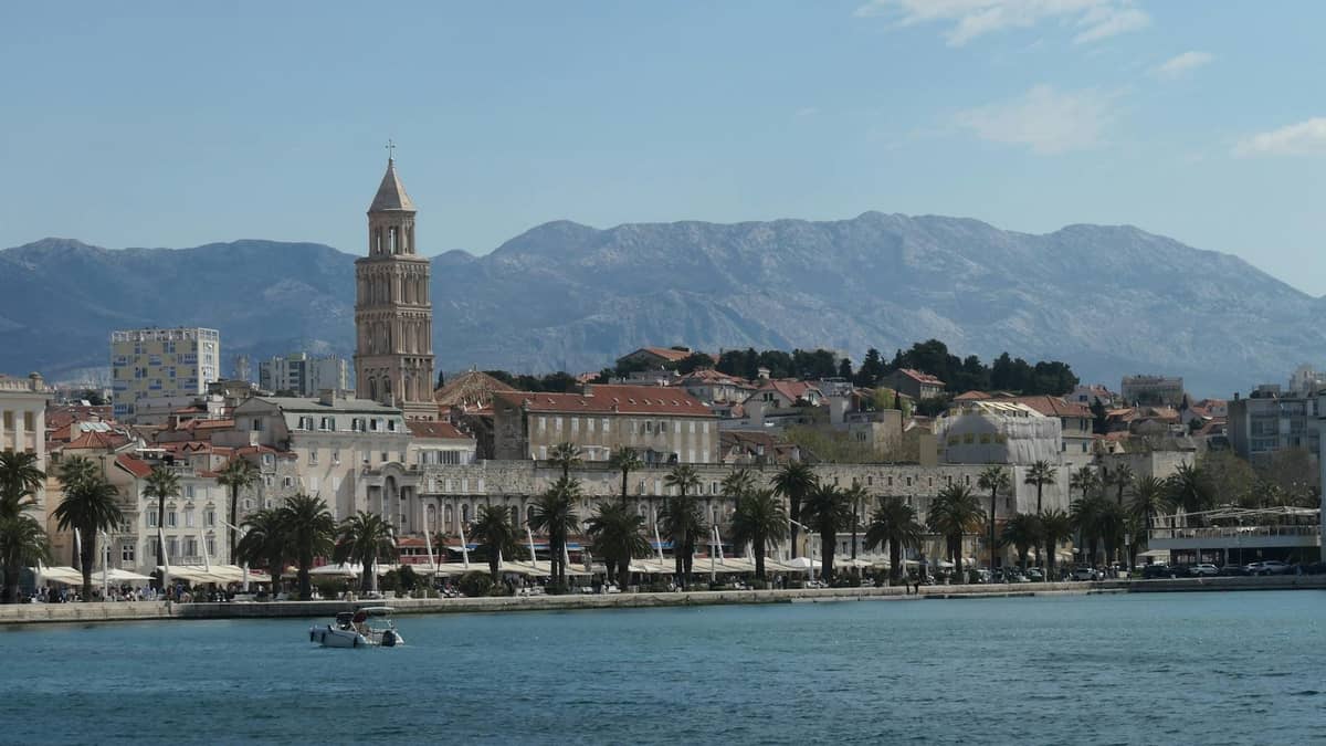 Panoramablick auf die Uferpromenade von Split, Kroatien, mit der Riva-Promenade, Palmen und einem Glockenturm vor Bergkulisse.