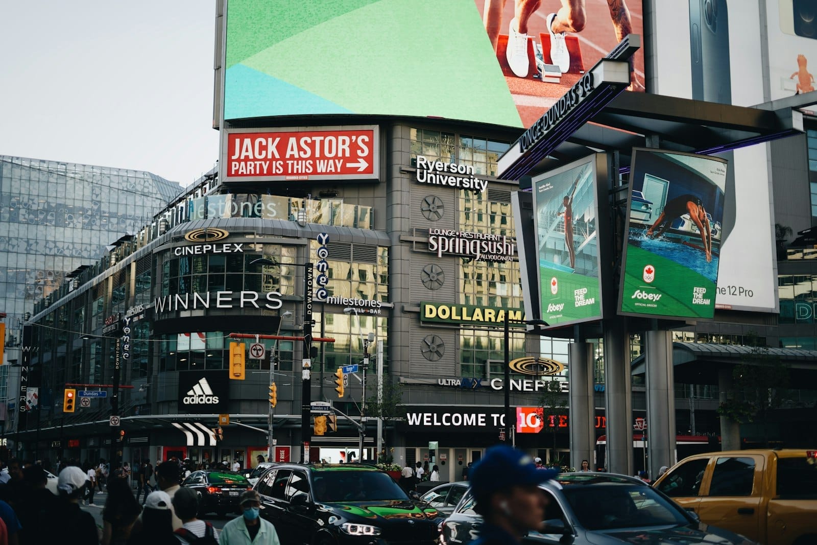 Movimentada intersecção no centro de Toronto em Yonge-Dundas Square, repleta de pessoas, carros e sinalização urbana reconhecível.