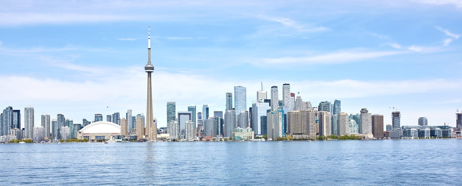 Panorama amplo e iluminado do horizonte de Toronto durante o dia, com a CN Tower e os arranha-céus refletidos na água sob um céu azul, transmitindo segurança e energia urbana acolhedora.