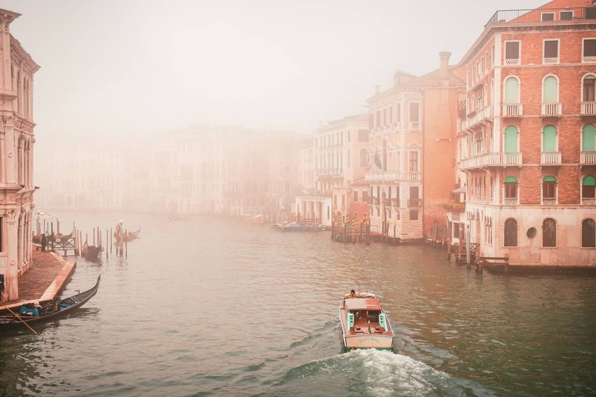 Venezianischer Kanal mit Booten und Gondeln, flankiert von bunten historischen Gebäuden in leichtem Nebel, der eine stimmungsvolle saisonale Szene schafft.