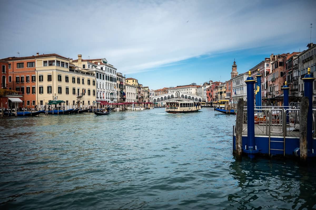 Panoramablick auf Venedigs Canal Grande mit der Rialtobrücke im Hintergrund, bunten historischen Gebäuden, Booten und blauem Himmel.