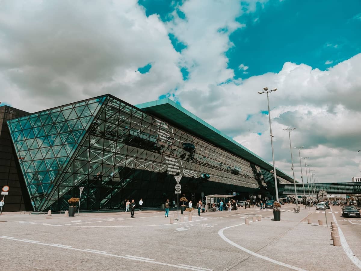 Façade en verre moderne de l'Aéroport Chopin de Varsovie avec des personnes et des véhicules devant sous un ciel partiellement nuageux.