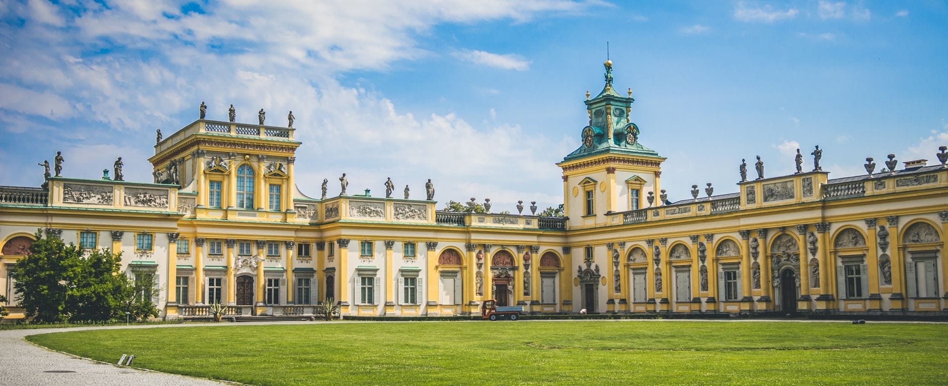 Vista ampla do Palácio de Wilanów em Varsóvia em um dia ensolarado, com céu azul e gramado verde à frente, exibindo uma arquitetura imponente.