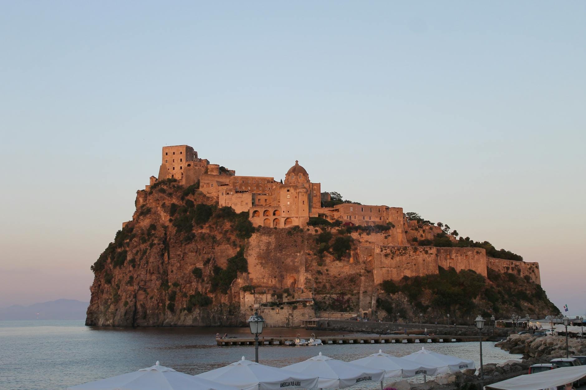 Il Castello Aragonese arroccato su un isolotto roccioso al largo della costa di Ischia al tramonto, con acque calme e un cielo dai tenui colori pastello sullo sfondo.