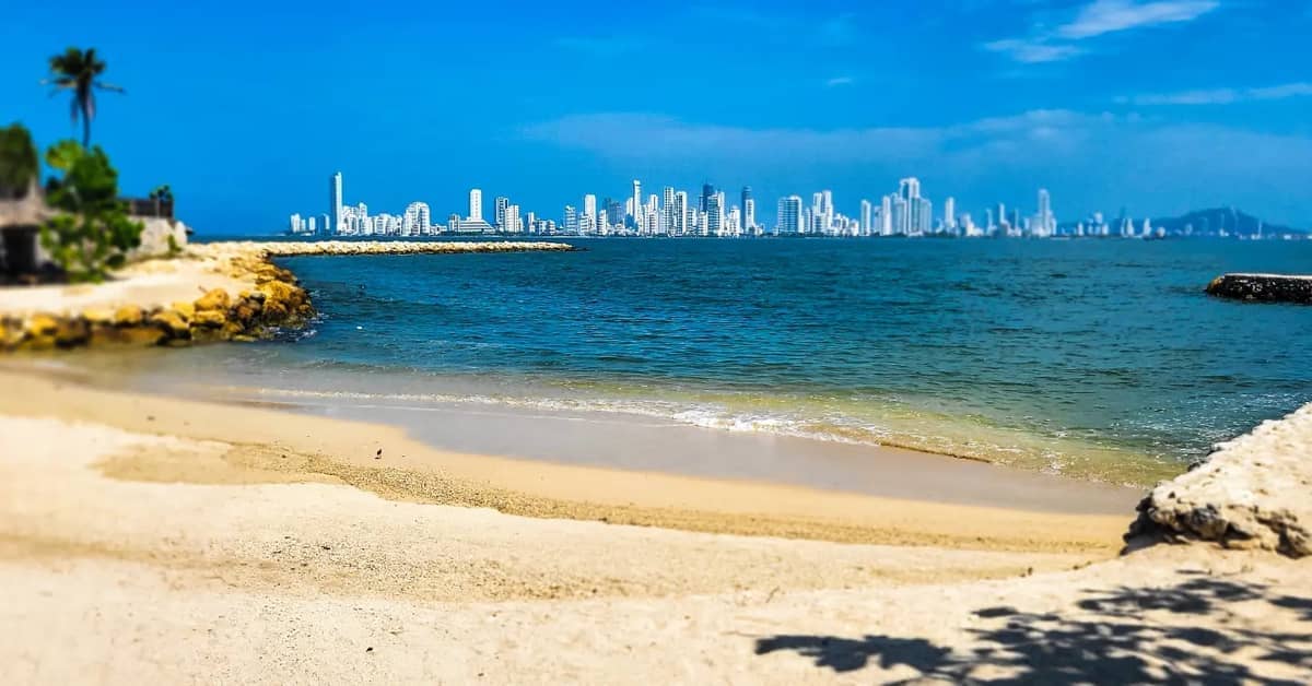 Sandy beach on Isla Tierra Bomba with calm turquoise water and Cartagena's modern city skyline visible across the bay under a clear blue sky.