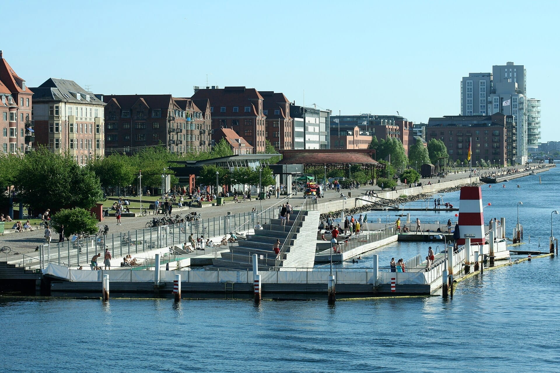 Vista dell'Islands Brygge Harbour Bath a Copenaghen con persone che godono delle piscine, del fiume e degli edifici urbani adiacenti in una giornata di sole.