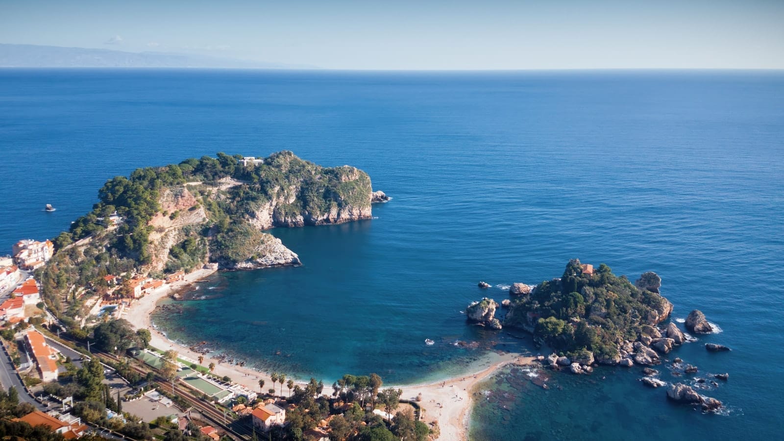 Luftaufnahme des Eilands Isola Bella, das durch einen schmalen Landstreifen mit der Küste Taorminas verbunden ist, umgeben von türkisfarbenem ionischem Wasser und sattgrünen Hügeln.