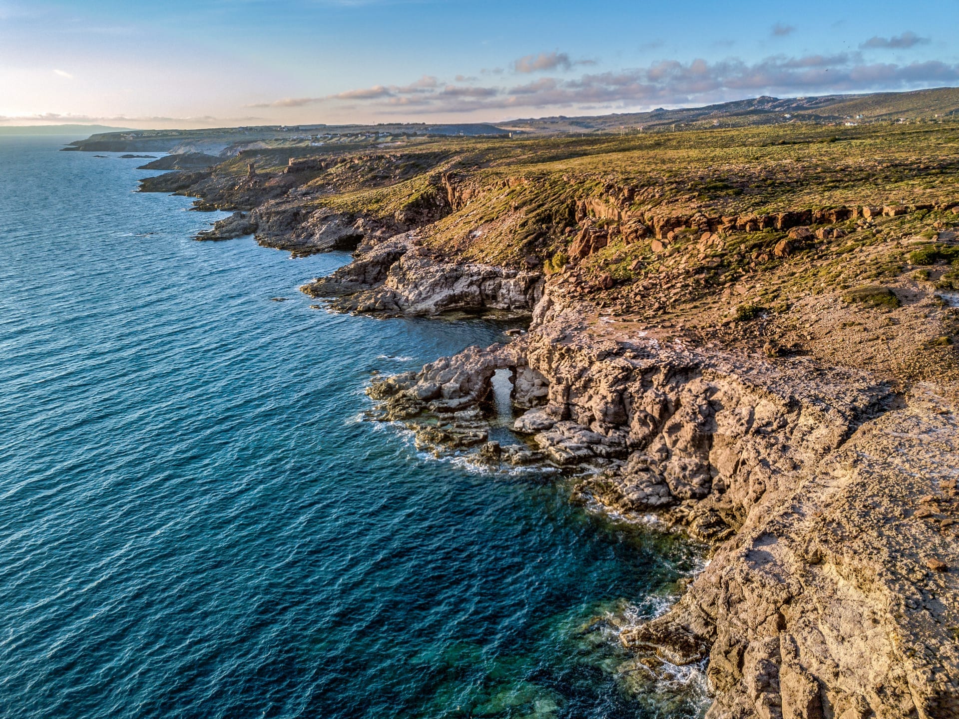 Felsige Küste der Isola di Sant'Antioco mit schroffen Klippen und klarem blauem Meer unter einem strahlenden Himmel zur goldenen Stunde.
