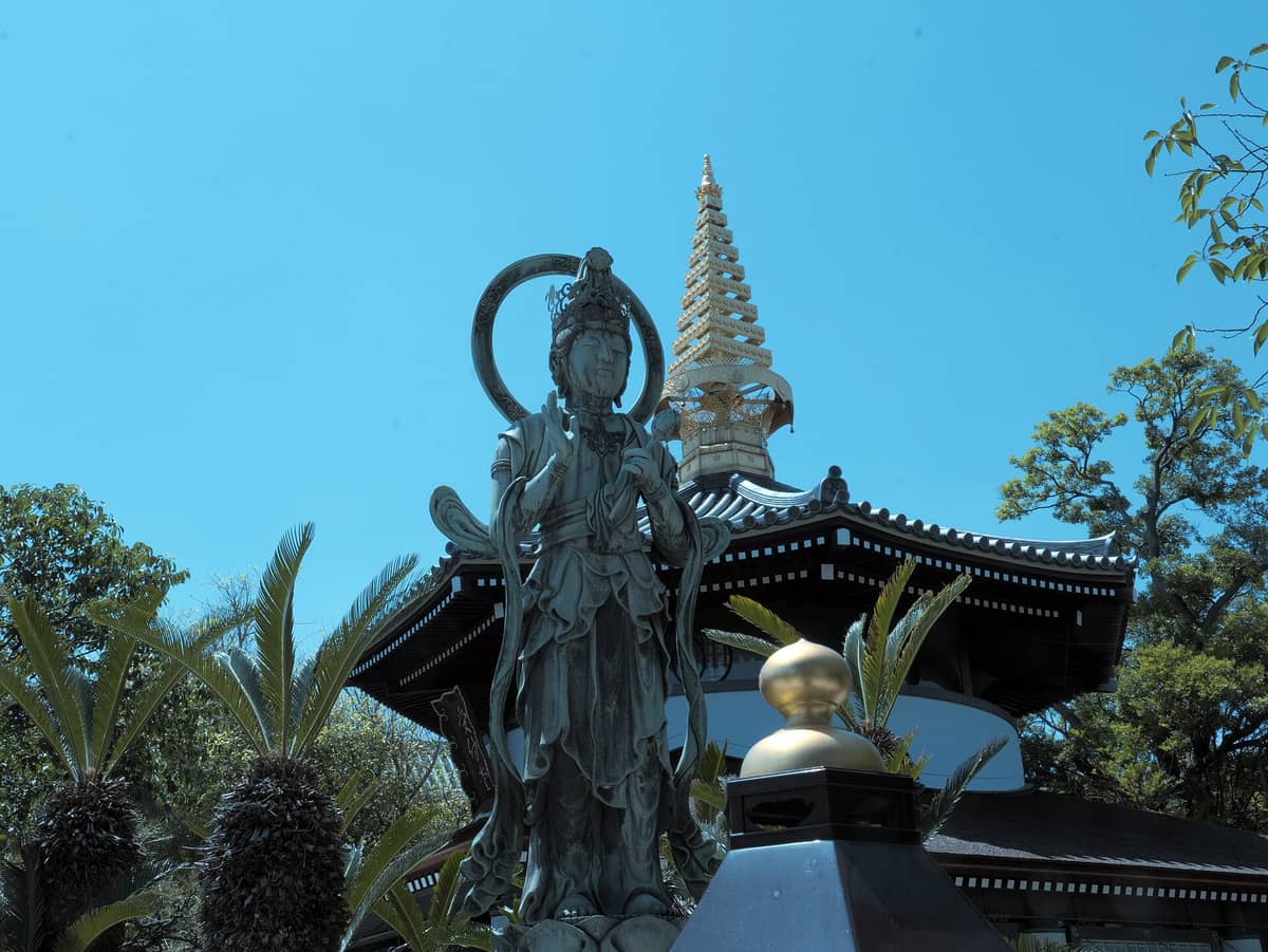 A statue of Kannon in front of the modern main hall and golden pagoda of Isshin-ji Temple in Osaka, under a clear blue sky.