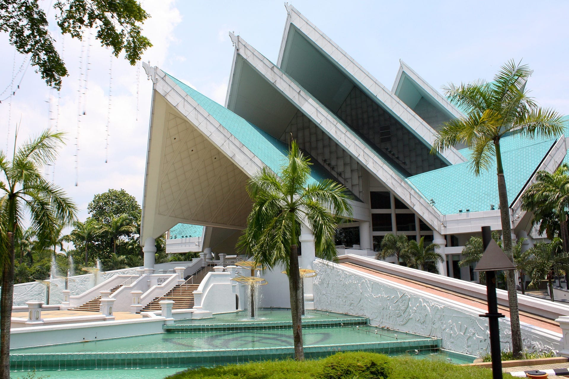 Istana Budaya’s striking modern architecture with angular turquoise roofs, white staircases, palm trees, and reflecting pools in bright daylight, Kuala Lumpur.