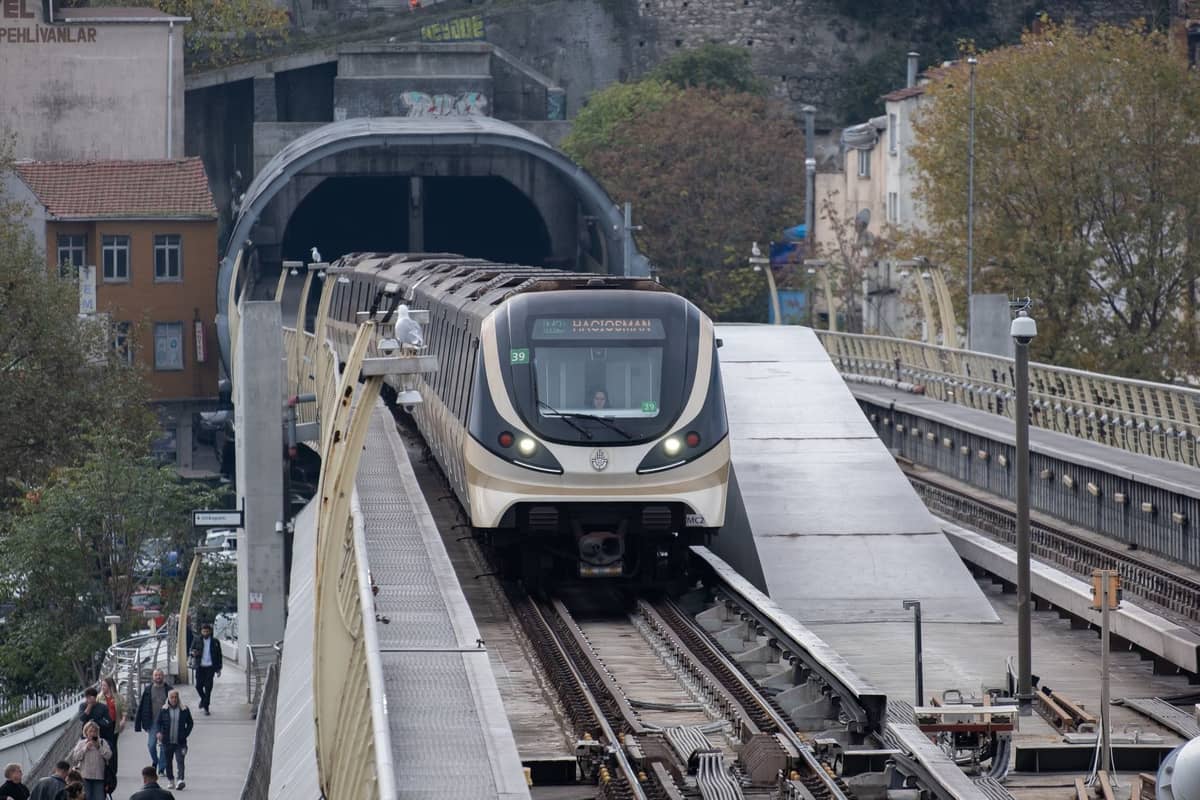 Istanbul-Metro-Zug überquert eine Brücke in Richtung eines Tunnels mit Stadtgebäuden im Hintergrund und Menschen, die in der Nähe spazieren.