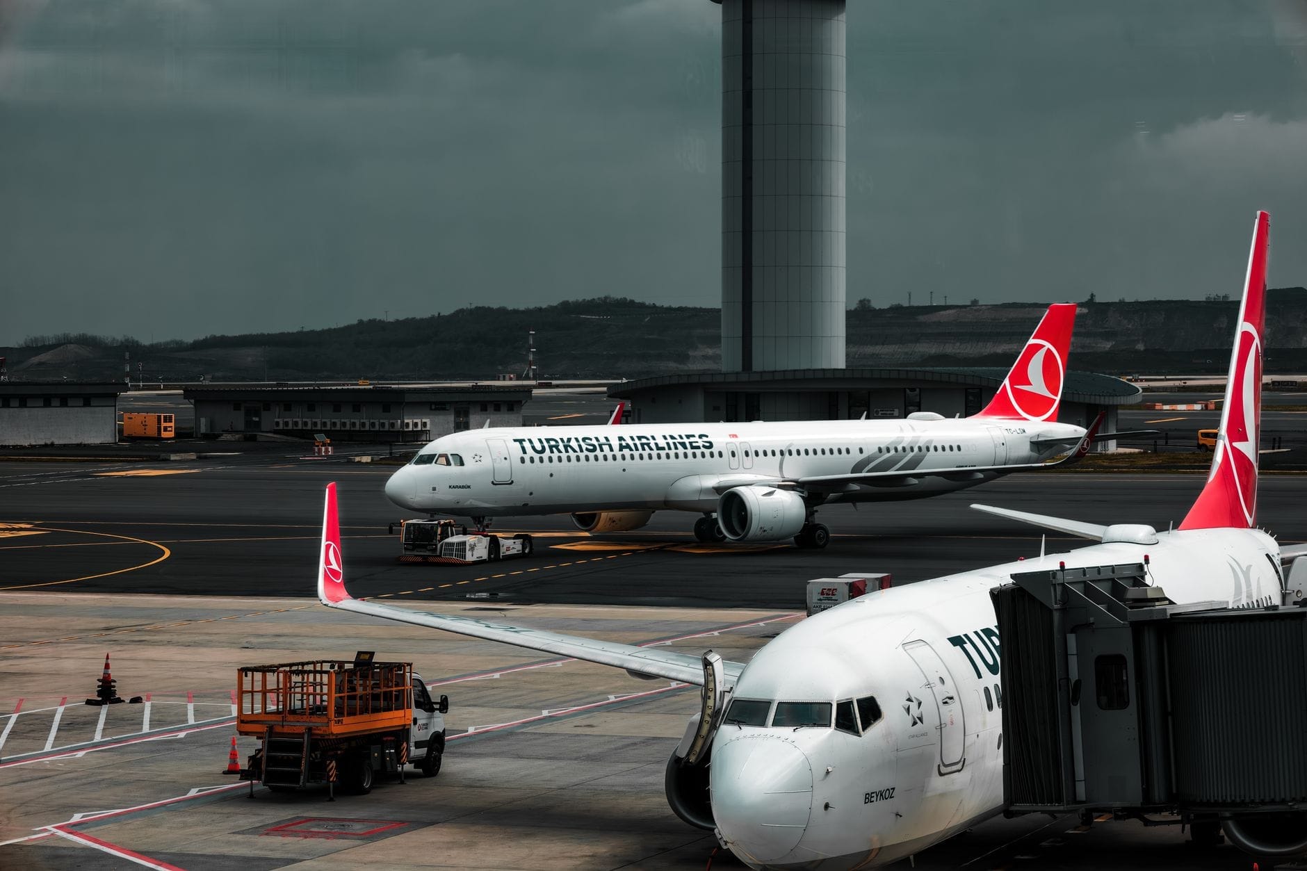 Aviões da Turkish Airlines nos portões e na pista do Aeroporto de Istambul, com o terminal e a torre de controle ao fundo sob um céu dramático.