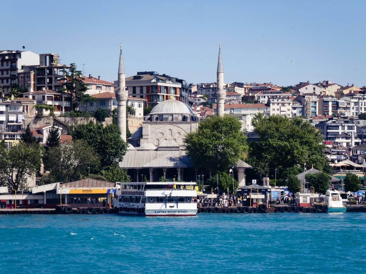 Набережная Üsküdar с историческою мечетью, паромами и жилыми домами на заднем плане в солнечный день.