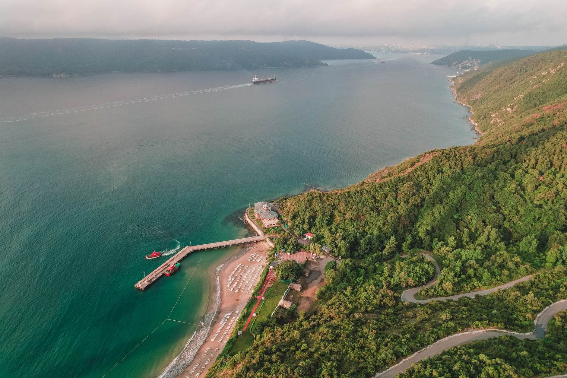 Vista aérea de um litoral cênico perto de Istambul com colinas verdes, uma praia de areia, um píer e um navio cargueiro ao longe sobre águas azuis calmas.
