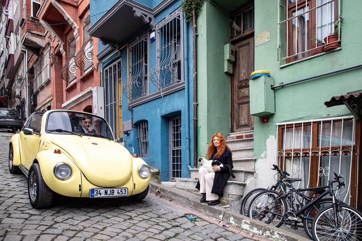 Woman sitting on stoop of colorful street in Istanbul petting a cat, with historic buildings and a yellow car nearby.